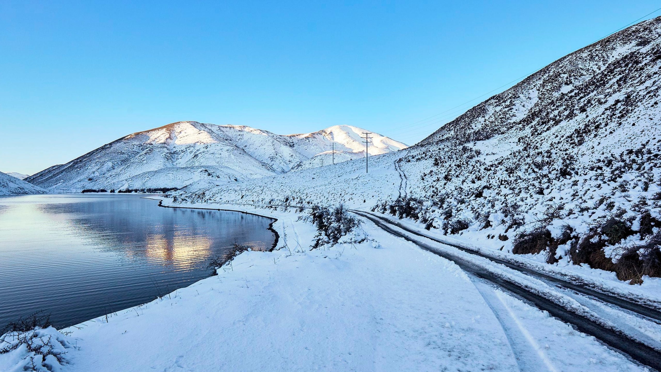 A snowy mountain landscape with a calm lake on the left, a winding road on the right, and snow-covered peaks under a clear blue sky.
